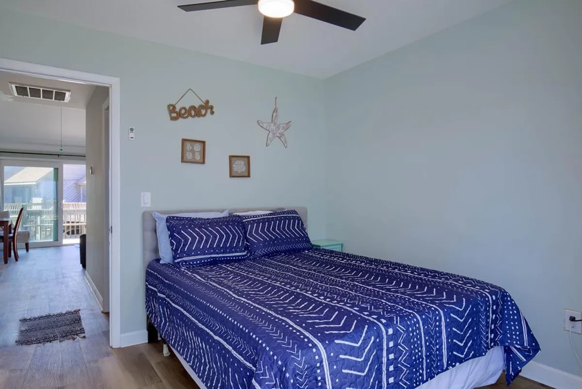 This image features a tranquil bedroom with a bed adorned in blue patterned bedding, a window letting in natural light with an outdoor view, and beach-themed wall decor. The setup communicates comfort and the refreshing atmosphere of a vacation stay.