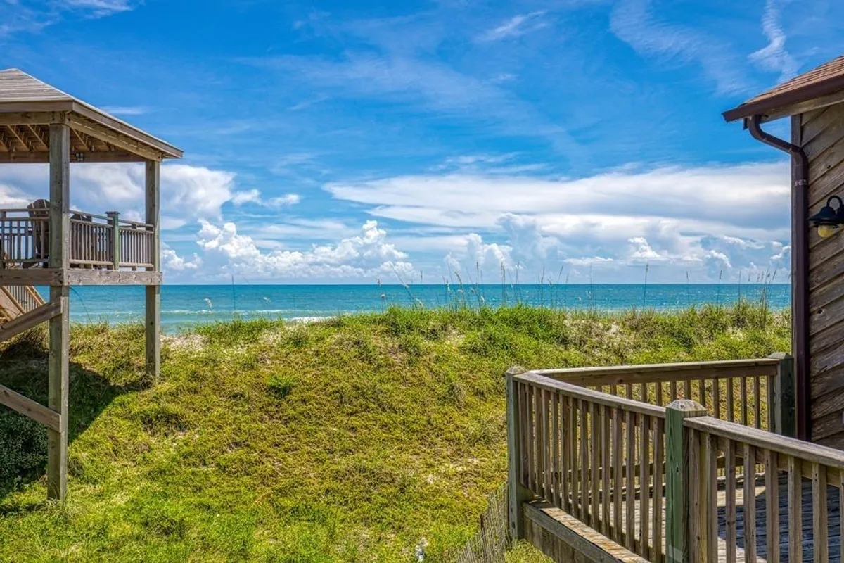 A wide view from a wooden deck overlooking dunes, the ocean, and a dramatic, cloudy blue sky. The space evokes relaxation and offers a clear sense of proximity to the beach, ideal for morning coffees, beach walks, or shared sunsets.