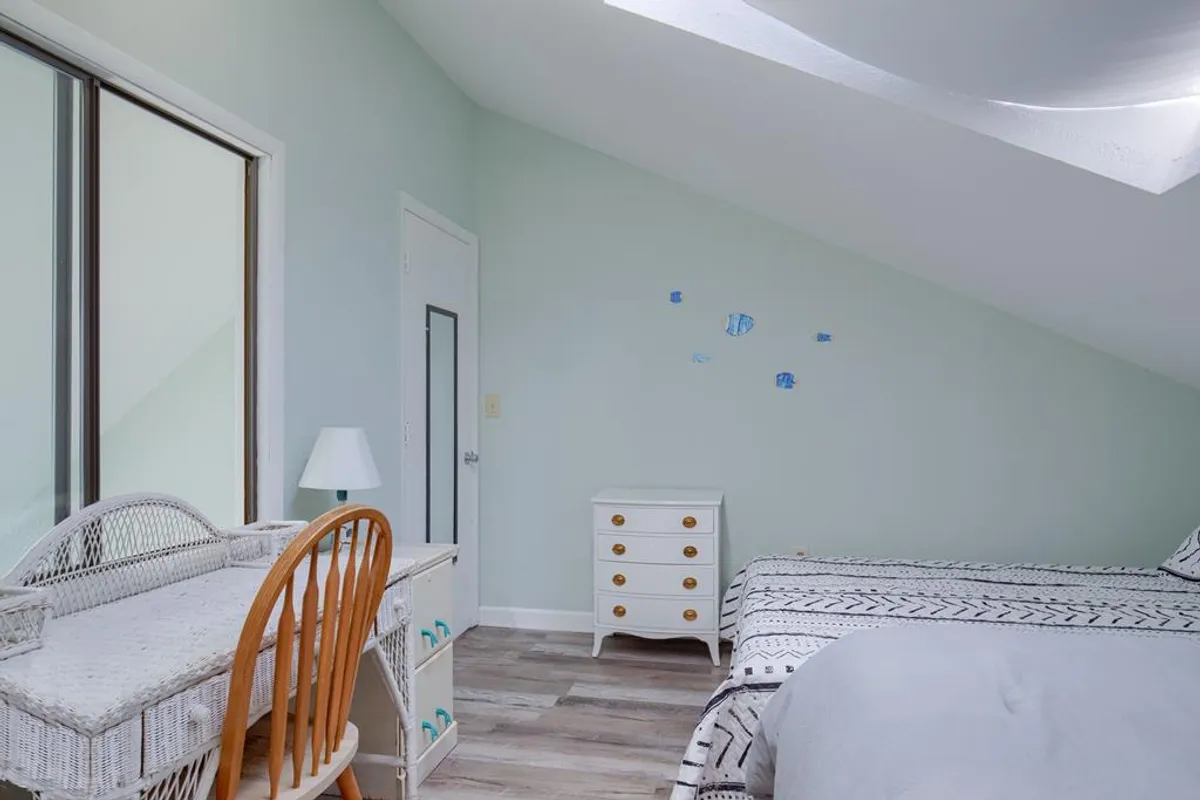 Inside, a serene bedroom features a wicker desk and chair, a white bedside table, and a bed with patterned bedding. The space is cozy and inviting, suggesting flexibility for both work and relaxation during a longer stay.