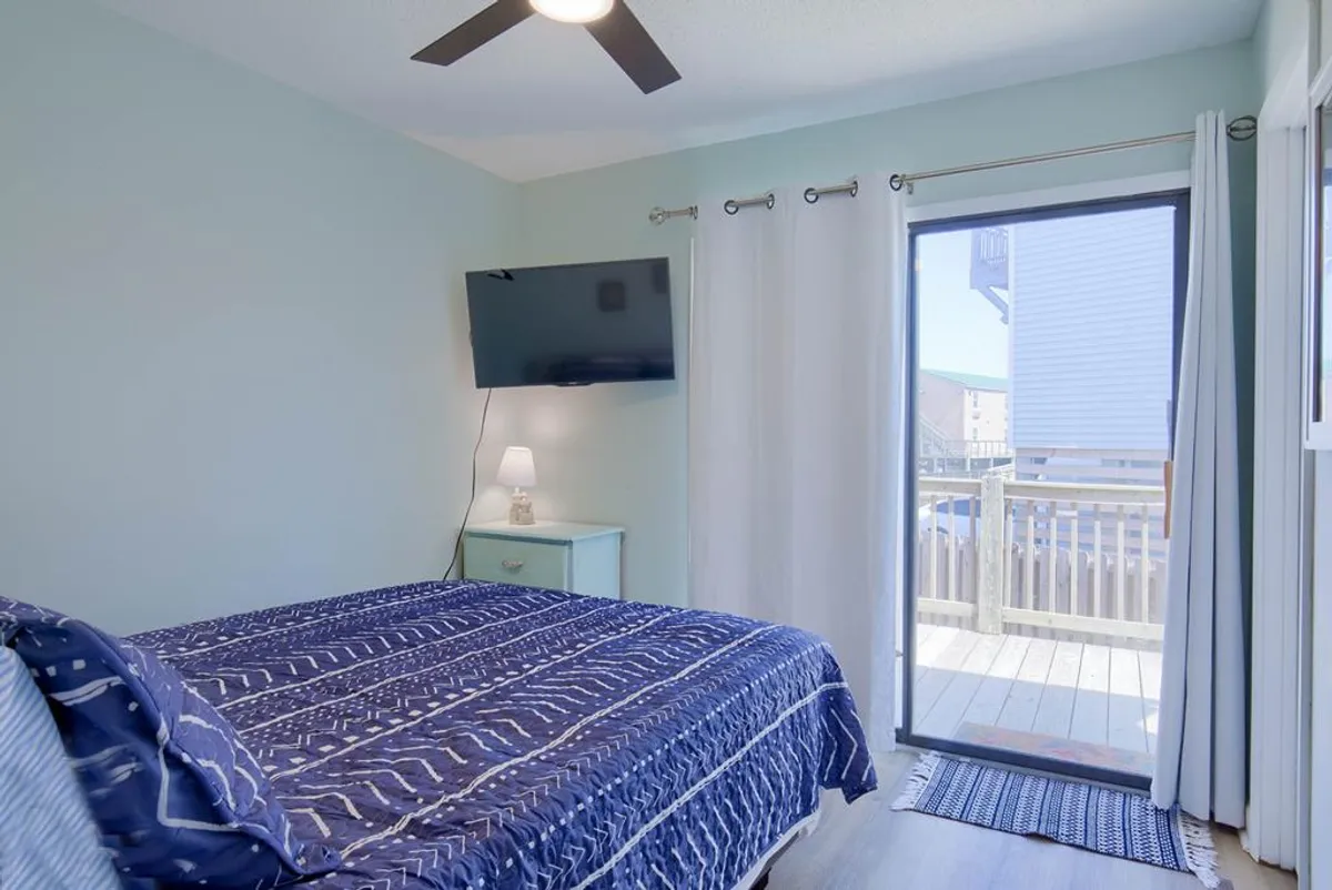 A tranquil bedroom featuring a bed with patterned bedding, a sliding glass door that opens to a sun deck, a TV mounted on the wall, and a nightstand with a lamp. Sunlight fills the room, creating an inviting haven for rest and relaxation.