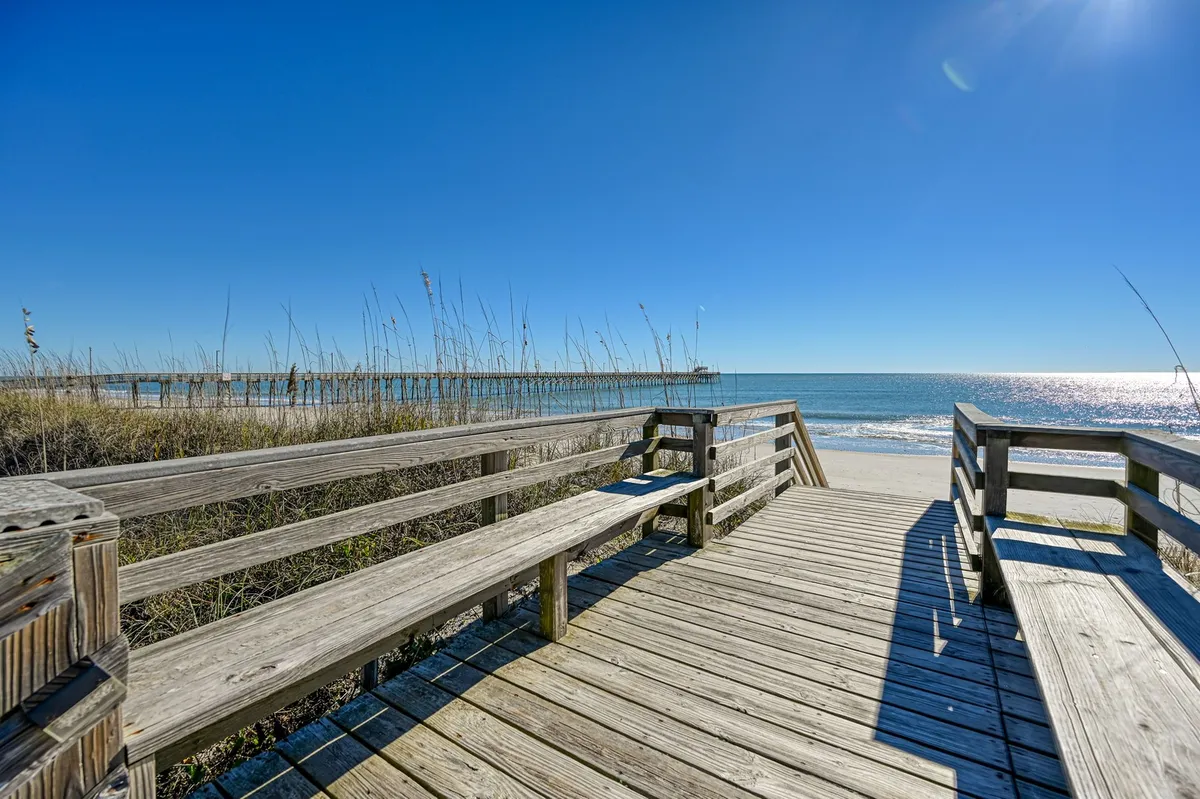 This exterior photo features a wooden boardwalk leading through dune grass toward the ocean, with the pier visible in the distance. The setting feels tranquil and immersive, offering a clear sense of how close and accessible the pier is from the property. The view highlights the natural beauty and direct path to beach activities and fishing.
