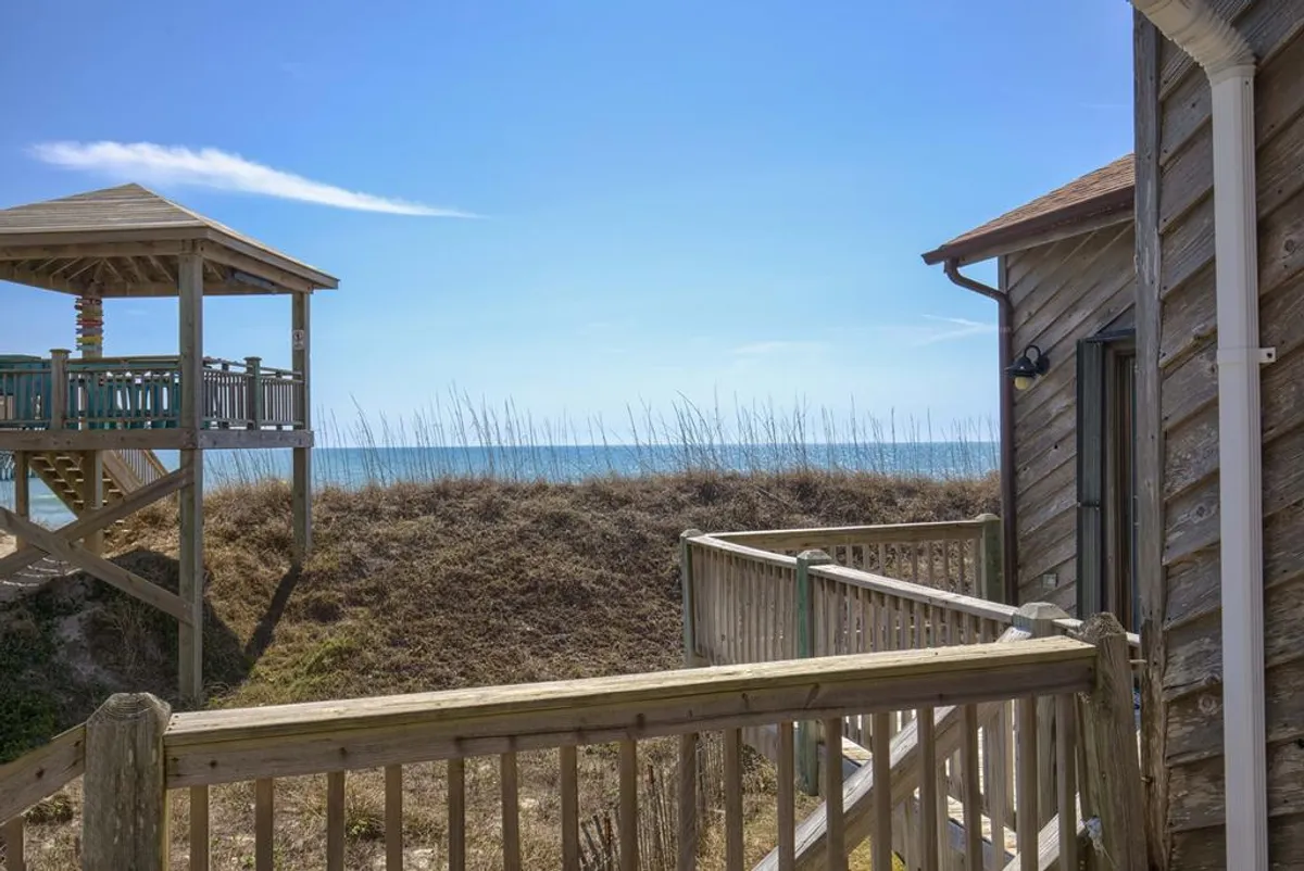 A scenic view of a wooden gazebo nestled amidst natural coastal vegetation with the ocean as a backdrop. The setting is peaceful and ideal for relaxing, sharing outdoor meals, or watching children play on the shore.