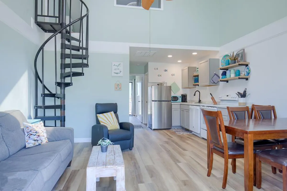 The image showcases a bright, inviting open-concept living and dining area featuring a cozy couch, a family-style dining table, and a striking spiral staircase leading to an upper loft. Sunlight streams in, illuminating the space and highlighting the comfortable, welcoming atmosphere ideal for relaxing, sharing meals, or gathering with loved ones.