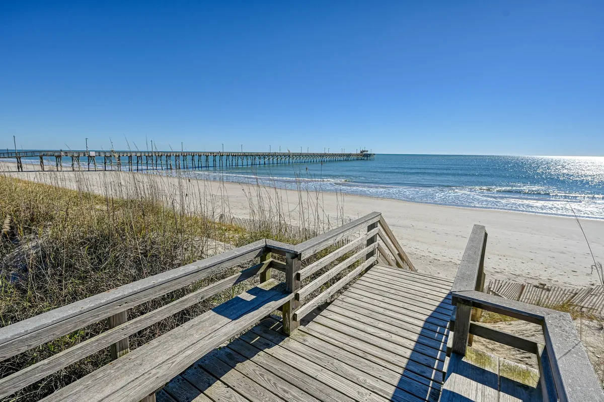 An inviting exterior view of a wooden boardwalk that leads over sandy dunes toward the ocean, with the Seaview Pier in the background. The scene exudes relaxation and easy access to the beach.