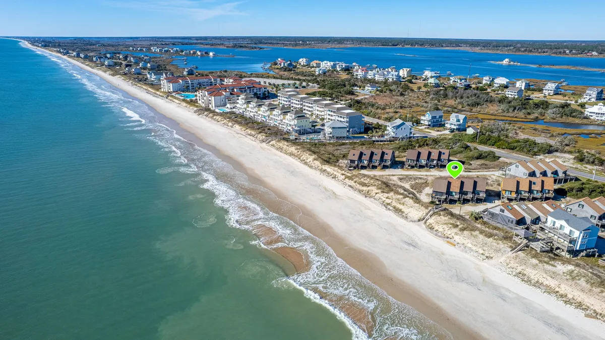 This wide-angle shot displays the shoreline with beach houses lining the coast, the ocean sparkling in the sunlight, and a hint of coastal activity. The view invites thoughts of beach outings, water sports, and a lively seaside atmosphere, with the pier and local amenities just a stroll away.