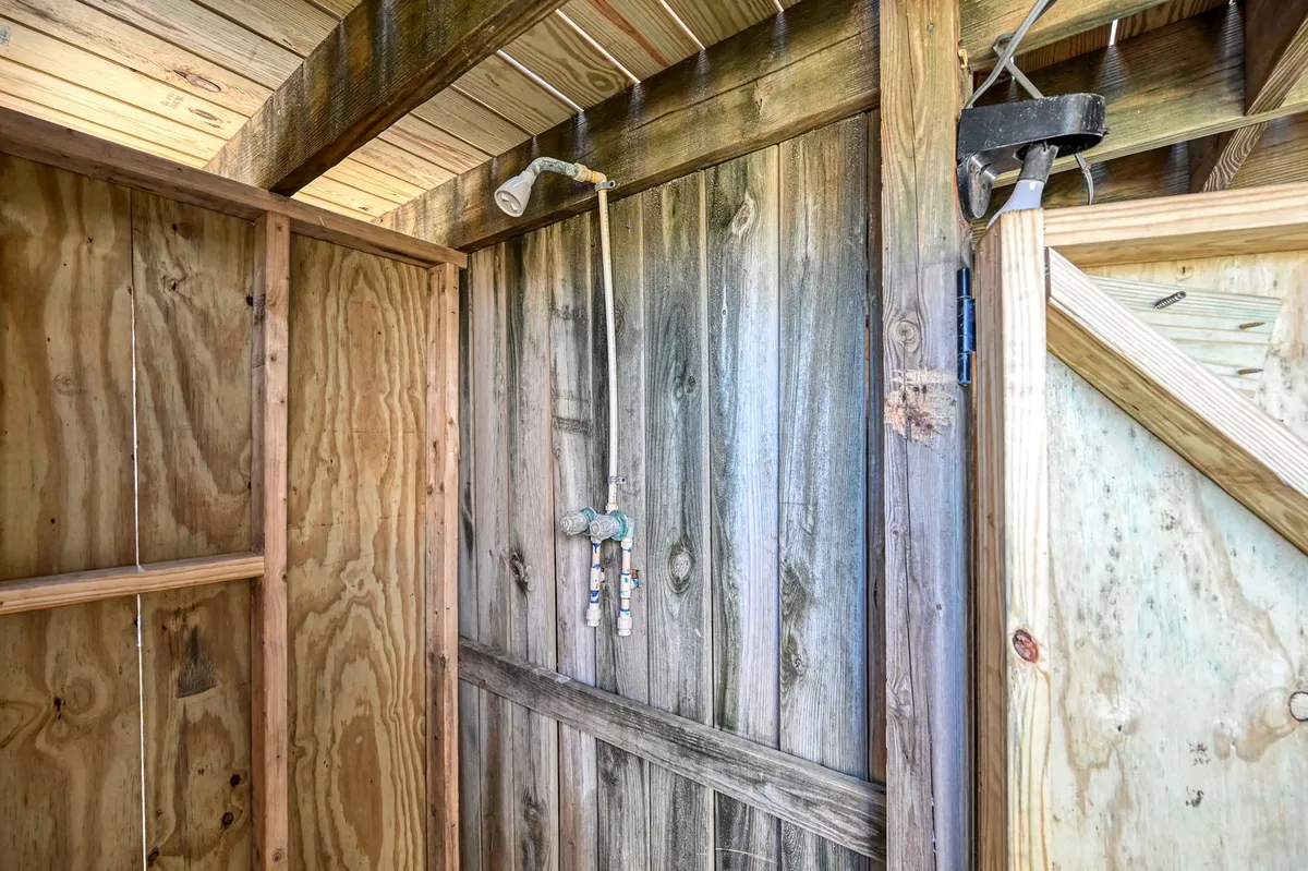 A rustic, open-air shower area featuring wooden walls, a shower fixture, and visible door hinges. The natural light and exposed elements emphasize the outdoor aspect, evoking the experience of rinsing off after a day at the beach.