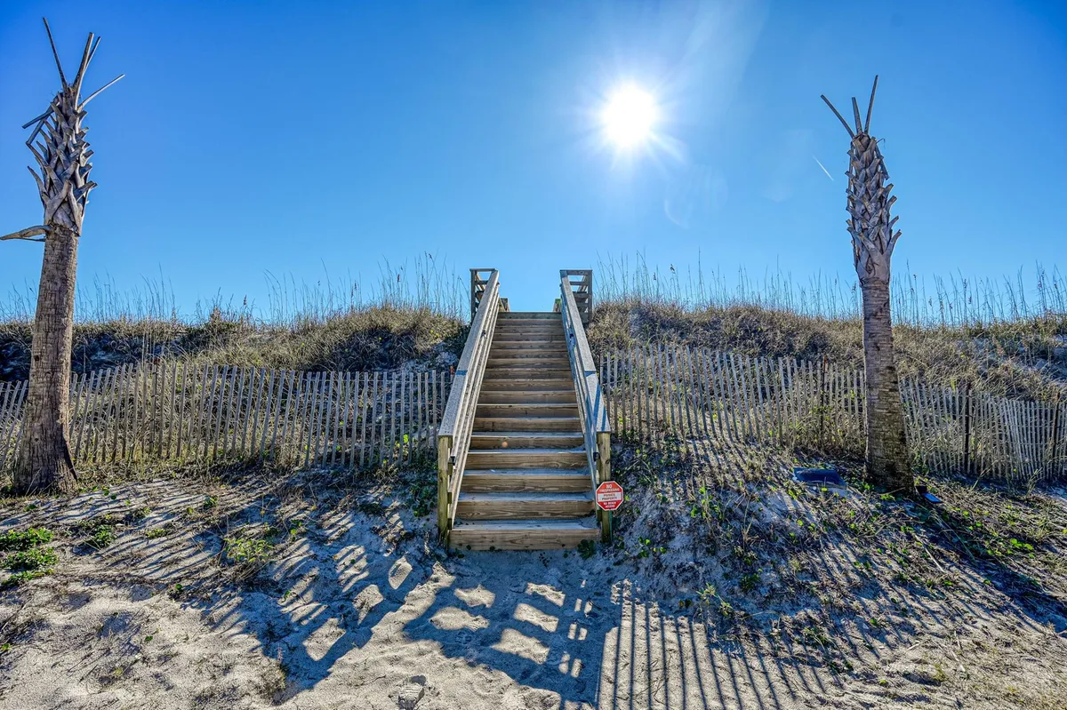 A sunlit view of wooden stairs bordered by palm trees and a fence, leading toward the beach. The scene conveys the inviting transition from the townhouse community to the shoreline.