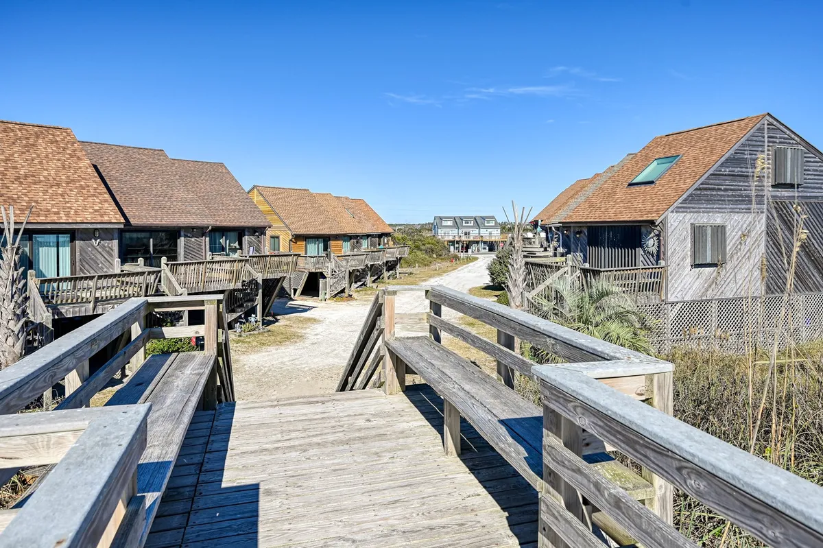 This exterior image features a wooden walkway leading toward a row of beach houses beneath a clear blue sky. The surroundings evoke a sense of tranquility, with easy access to the beach and the promise of outdoor relaxation. The scene references activities like walking to the beach, exploring the neighborhood, and enjoying nature.